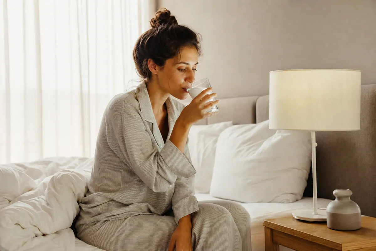 Personne qui se réveille dans son lit le matin et boit un verre d'eau posé sur la table de chevet