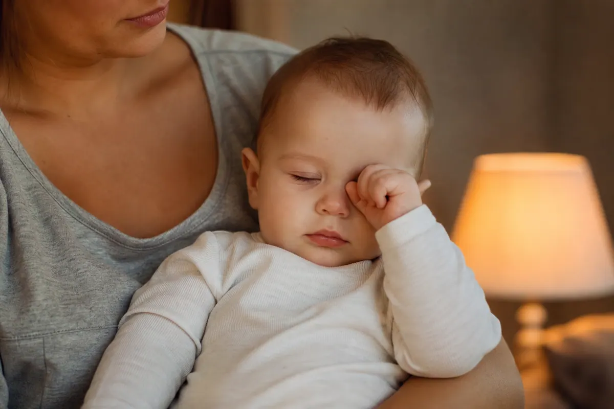 Bébé somnolent se frottant les yeux avec sa main dans les bras d'un parent, lumière douce du soir